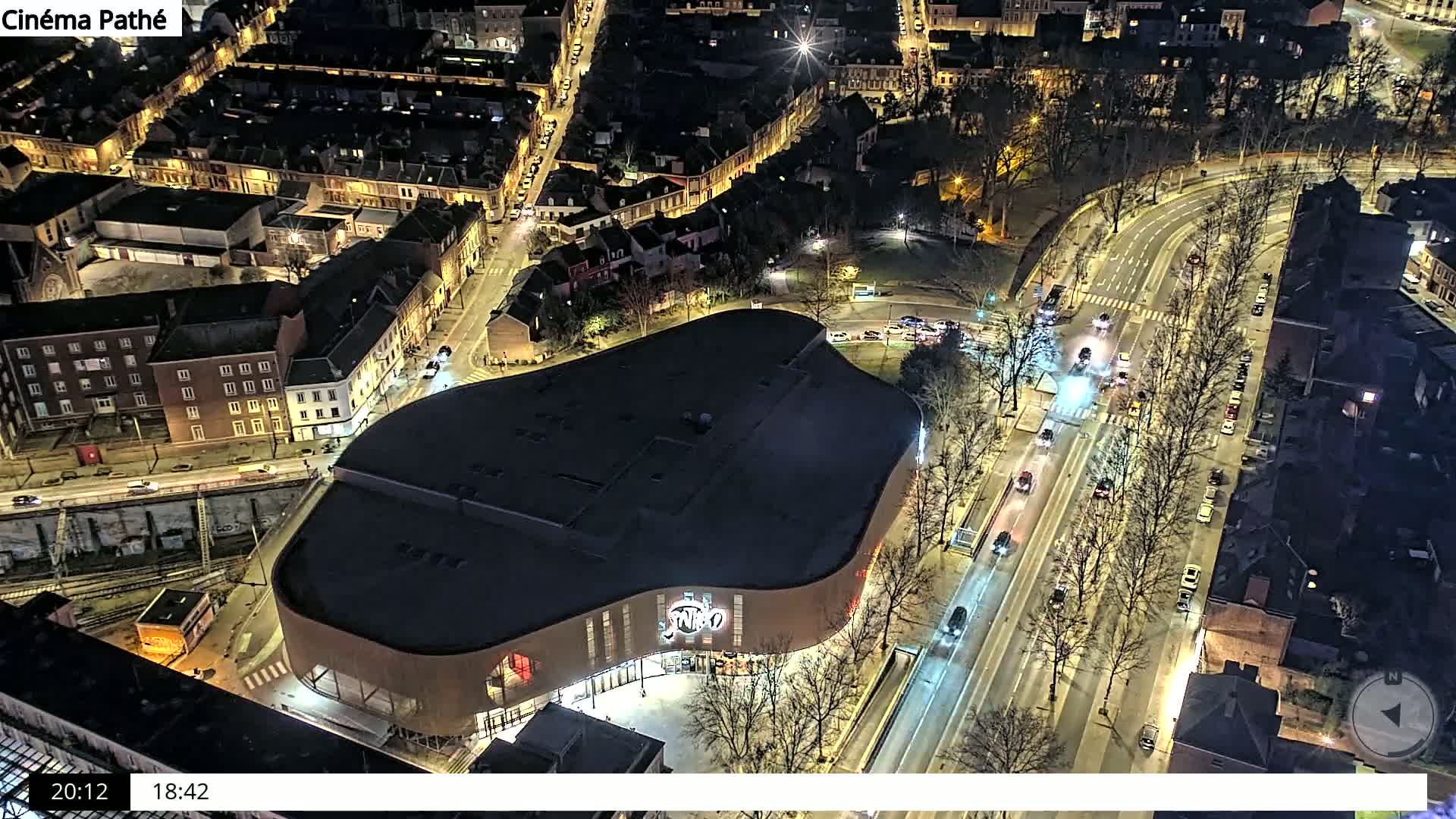 A large, dark-roofed building sits at the intersection of a busy, well-lit city street at night, surrounded by smaller buildings and trees.