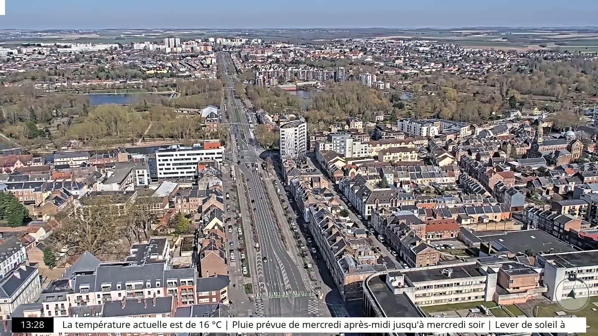 An aerial view of a city on a sunny day shows a long road cutting through residential neighborhoods and a park.