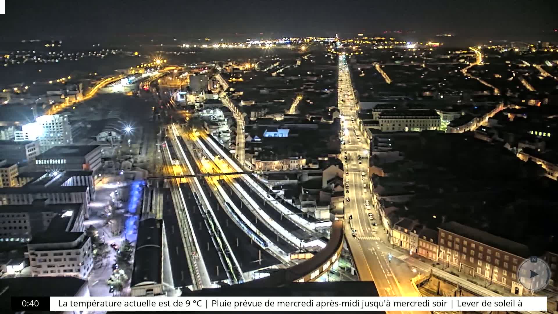 A nighttime aerial view shows a city's illuminated train station and streets.