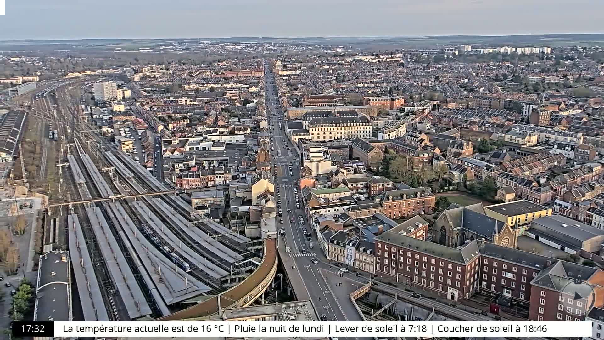 An aerial view of a city with a large train station, a main road, and numerous residential buildings under a partly cloudy sky.