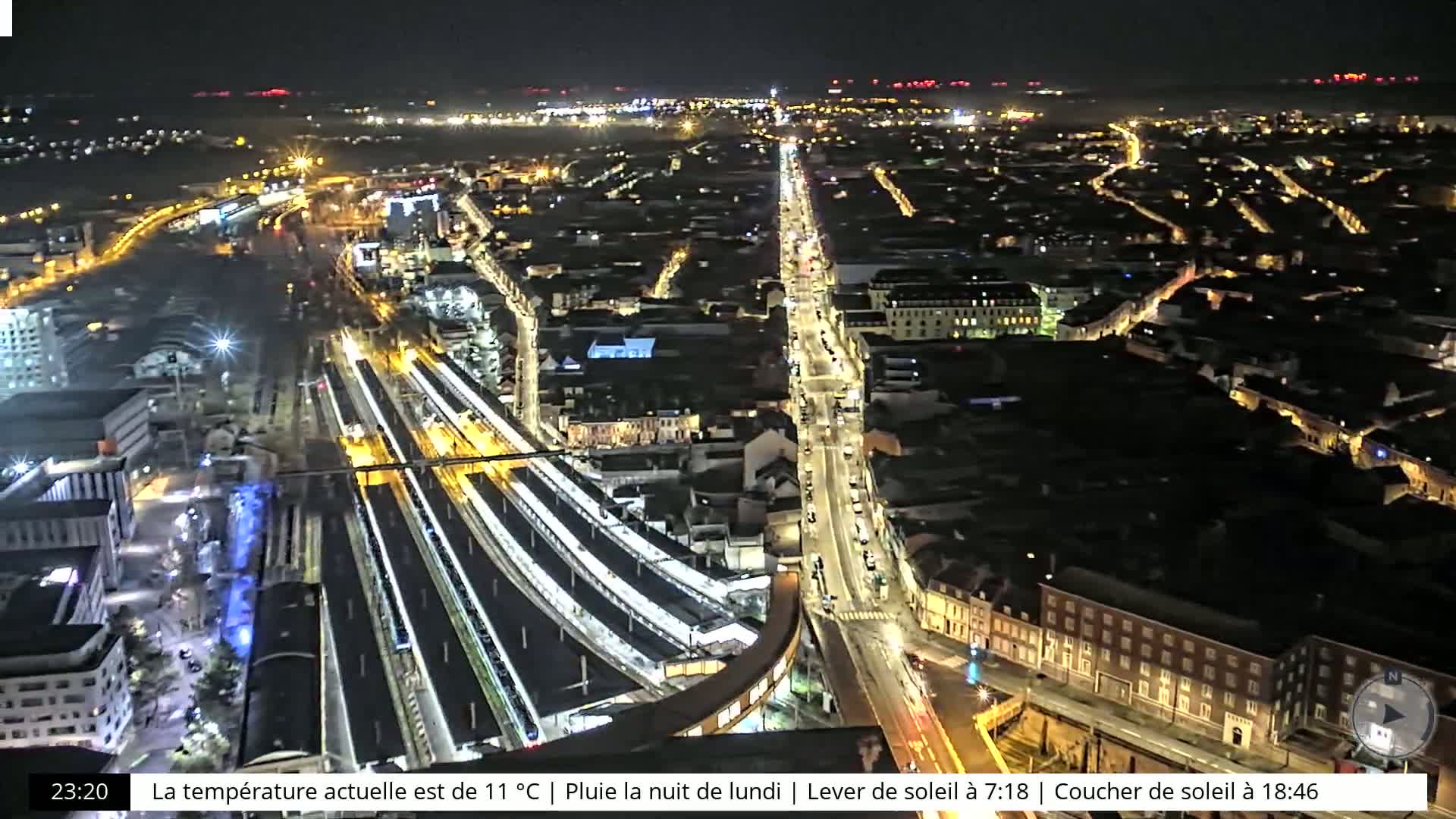 A nighttime aerial view of a city with a train station and illuminated streets, under rainy conditions.