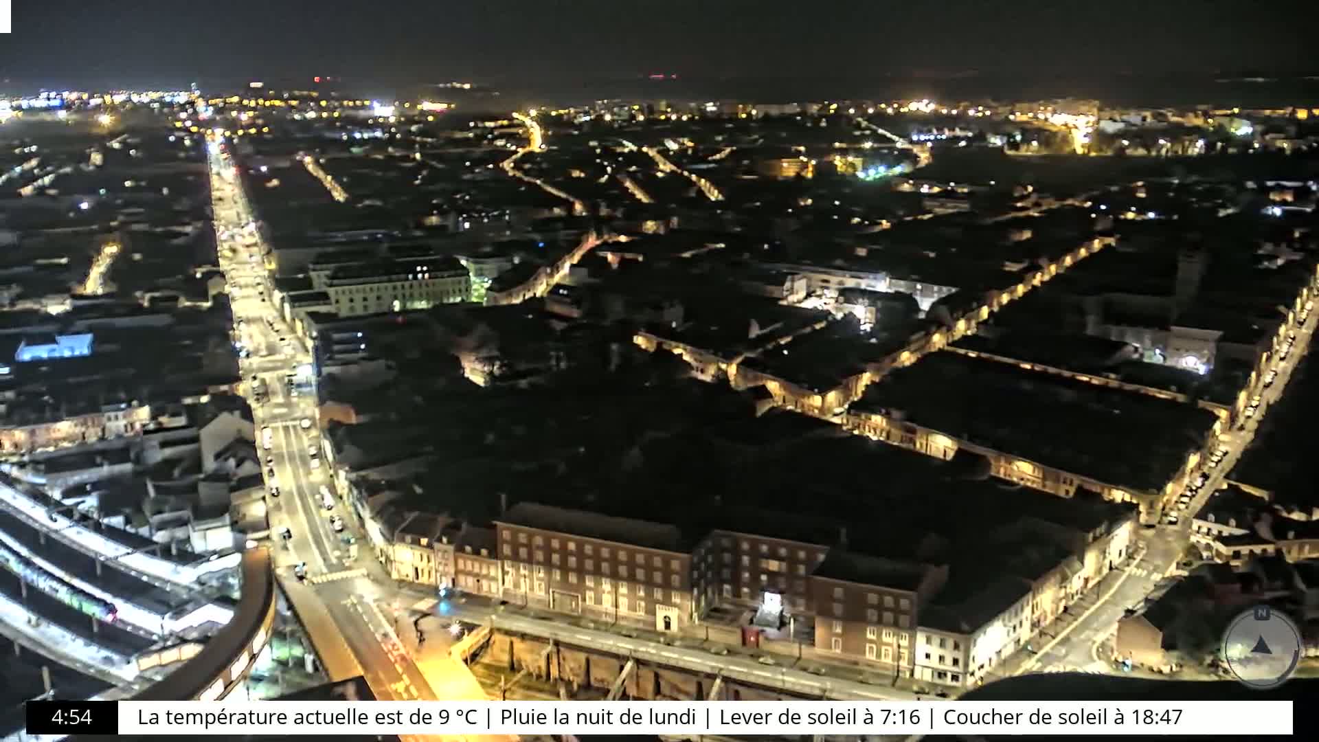A nighttime aerial view of a city with illuminated streets and buildings under rainy conditions.