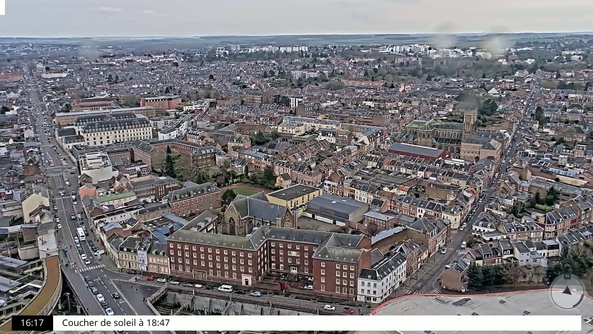 An aerial view shows a dense city of predominantly brick buildings under an overcast sky.