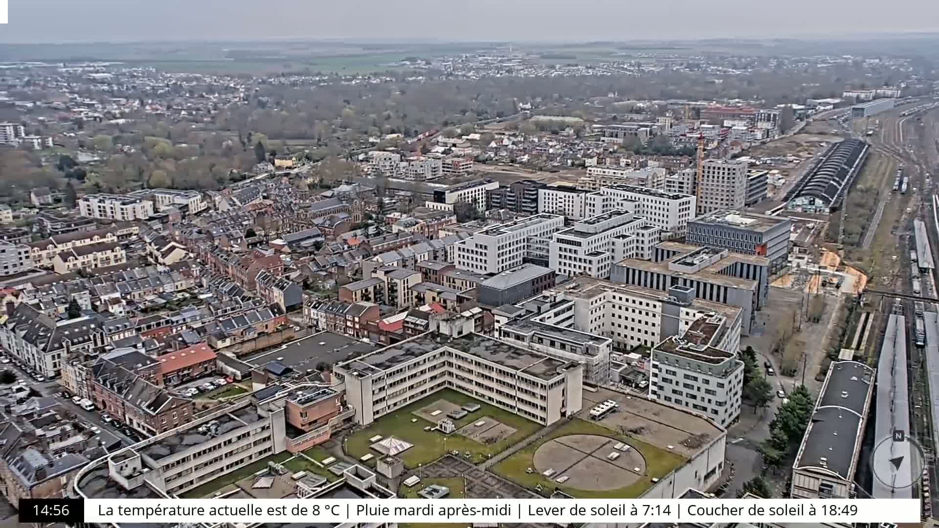 An aerial view of a city on an overcast day shows numerous buildings, including large apartment complexes, and a train station.