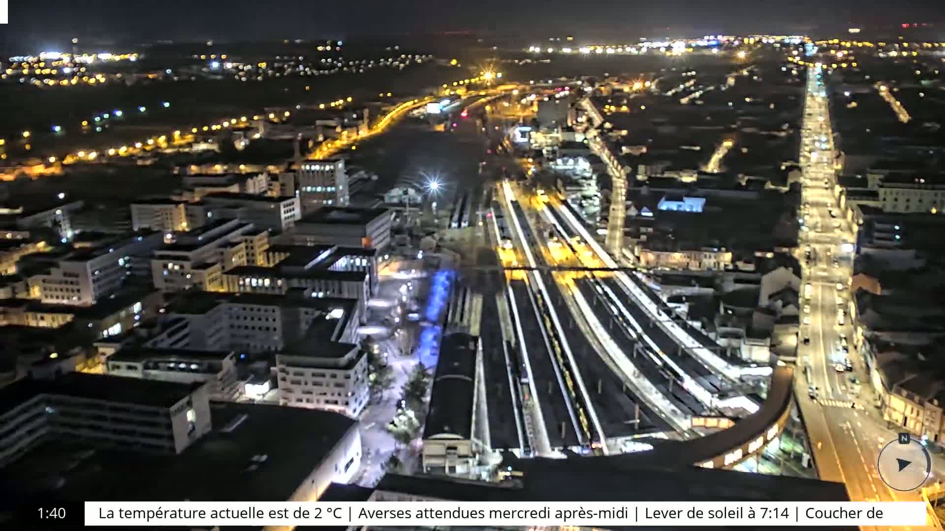A nighttime aerial view of a city with brightly lit streets and a train station, showing many buildings and roadways.