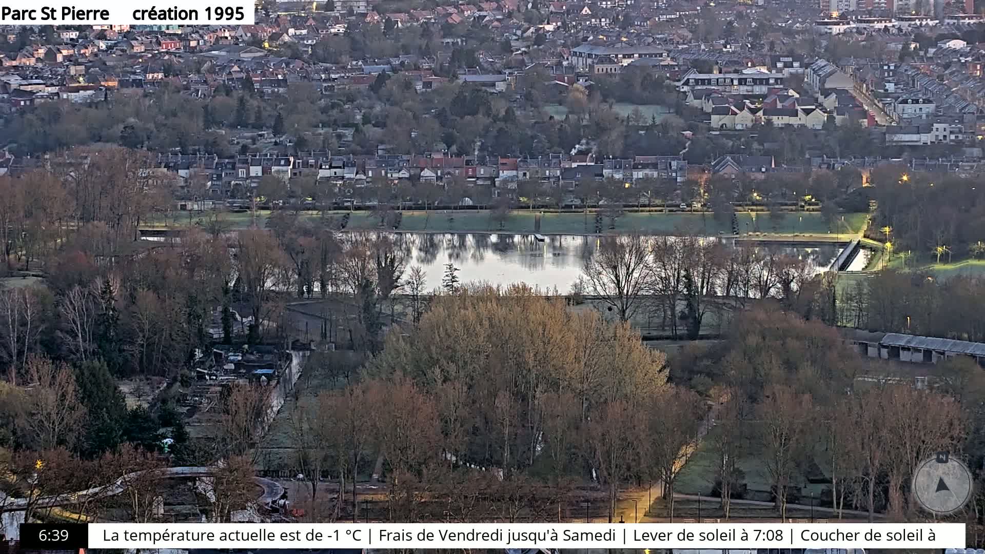 A high-angle, long shot of a park with a calm, reflective pond, surrounded by bare trees and residential buildings under a twilight sky.