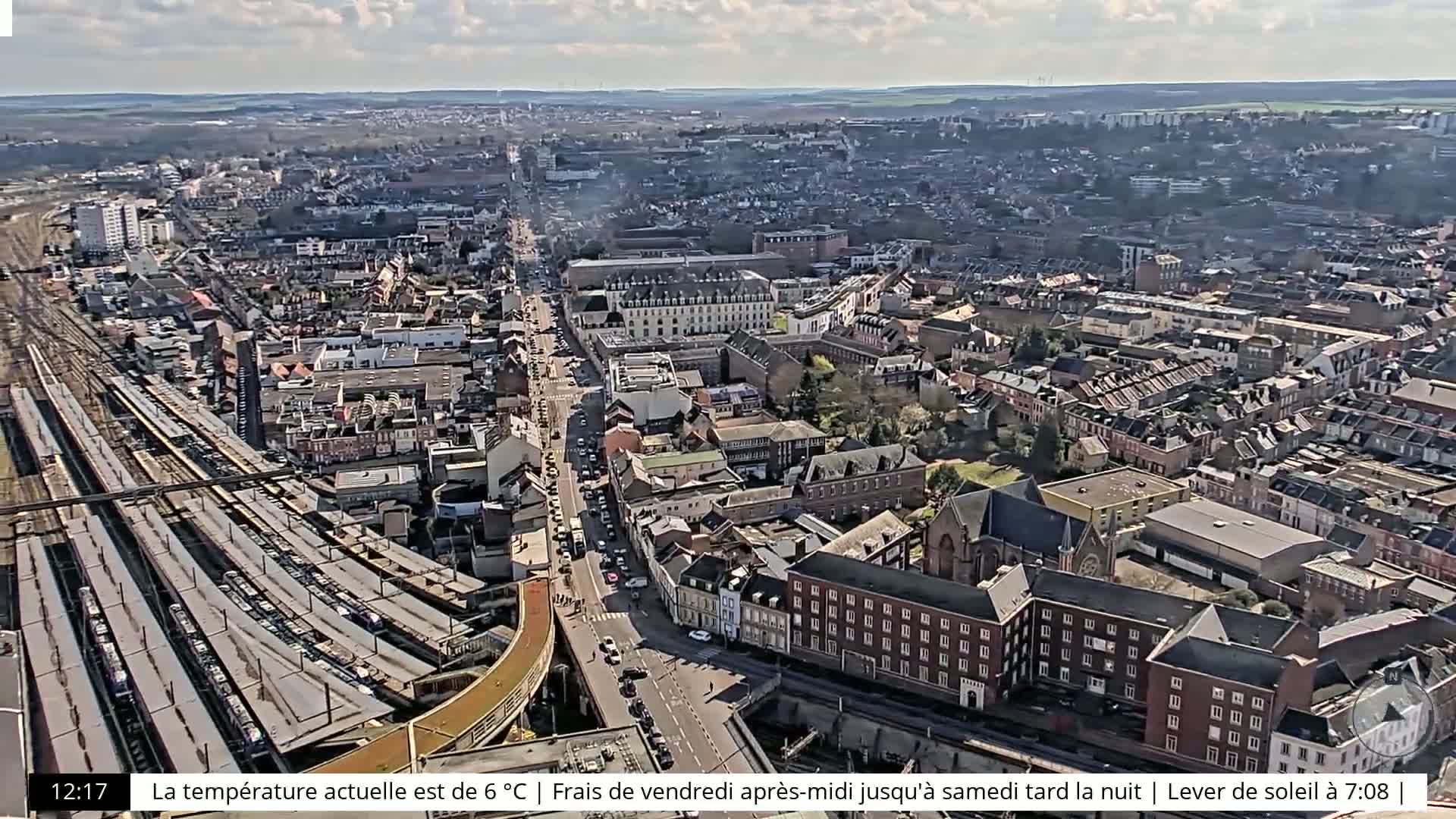 An aerial view of a city on a partly cloudy day shows a train station with many trains, roads with cars, and a variety of buildings of different sizes and styles.