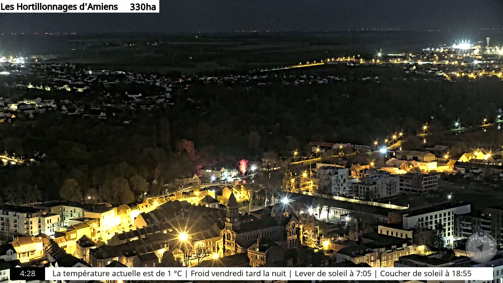 A nighttime aerial view of a city with illuminated buildings and streets, surrounded by dark trees and fields.