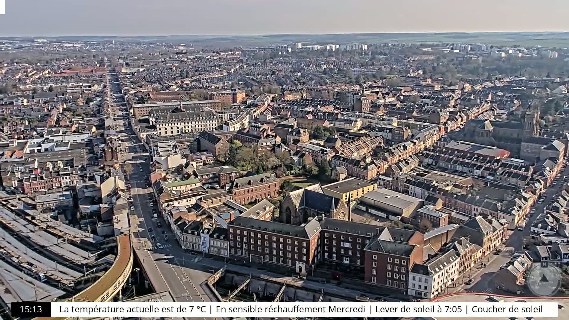 An aerial view shows a sprawling city on a sunny day, with a major road and train tracks visible.