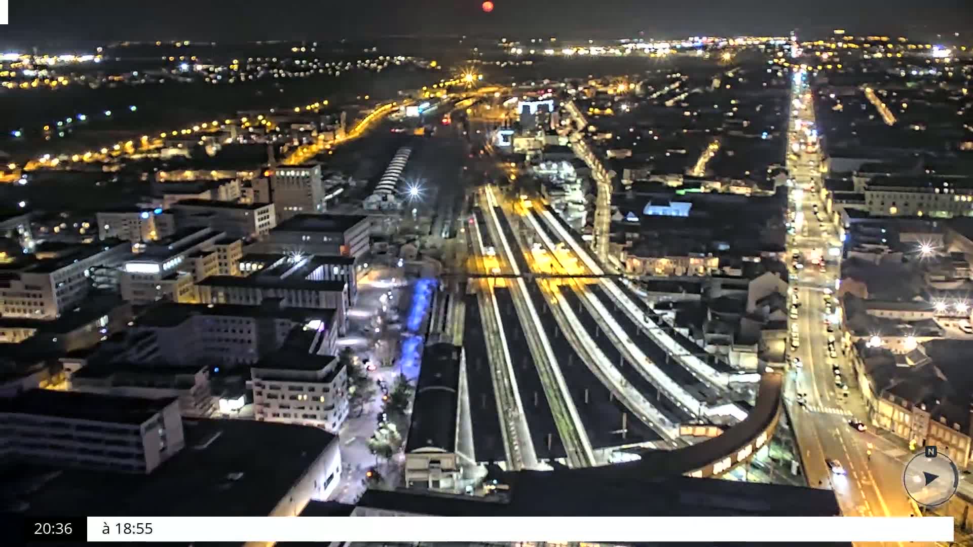 A nighttime, high-angle view of a city's illuminated train station and surrounding buildings, with a red moon visible in the distance.