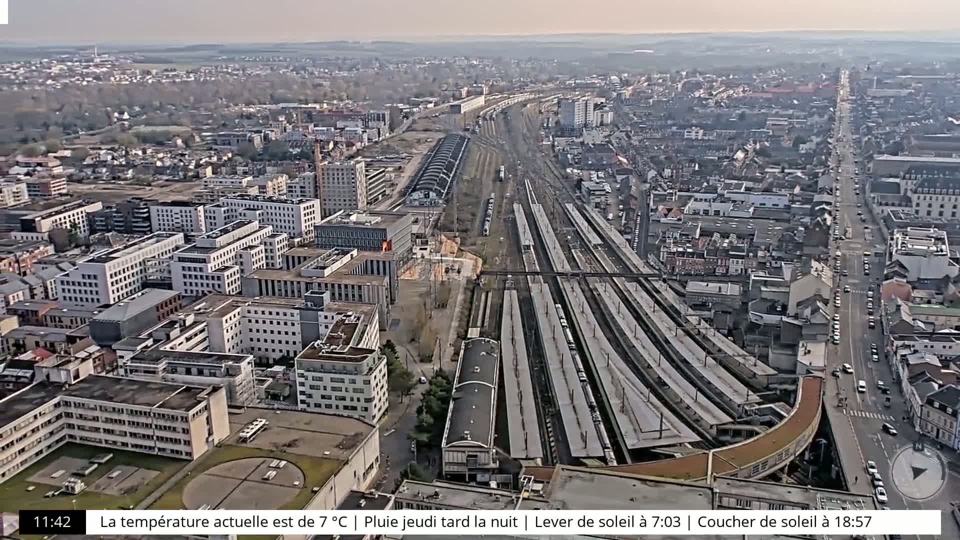 An aerial view of a city on a partly cloudy day shows a large train station with multiple tracks and several trains, surrounded by buildings and streets.