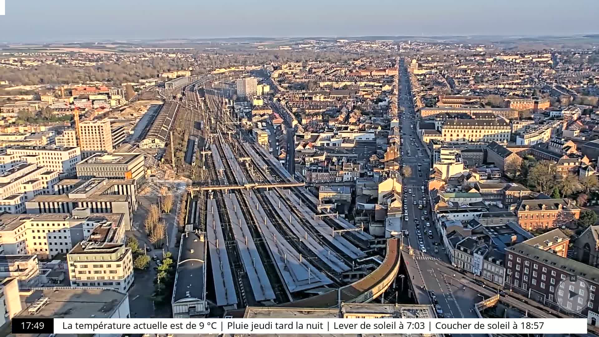 An aerial view shows a city with a large train station and a long road, under partly sunny conditions.