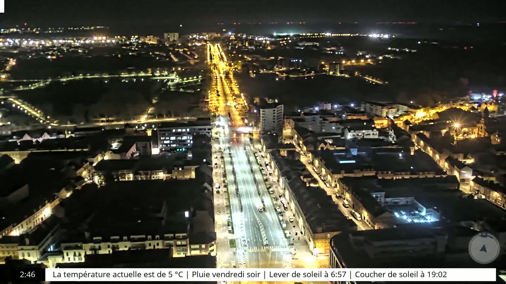 A nighttime aerial view of a city with a long, brightly lit main road, surrounded by buildings and dark parkland.