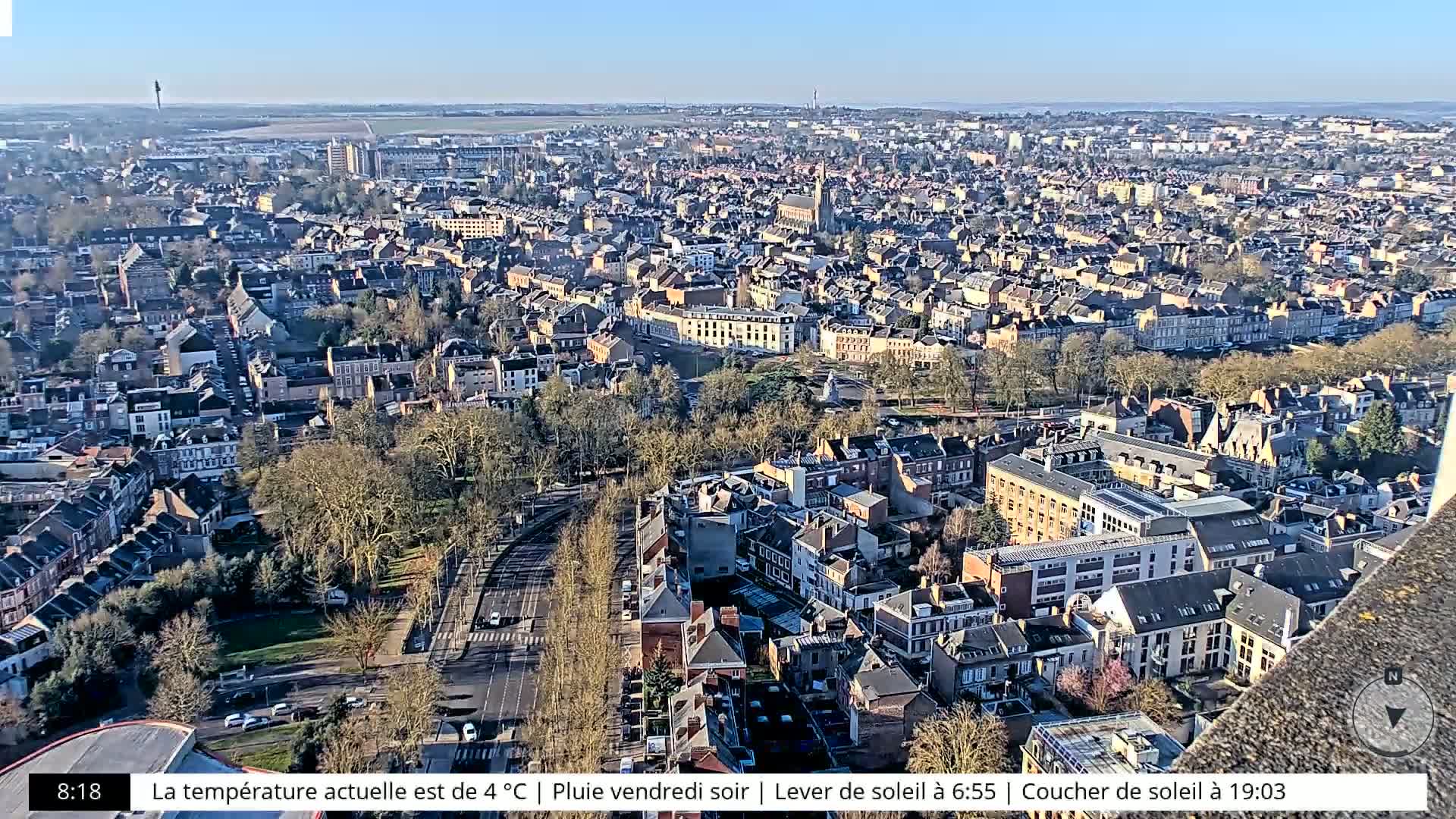 A high-angle, long shot of a city on a sunny day shows many buildings and a large park, with a clear sky visible in the background.