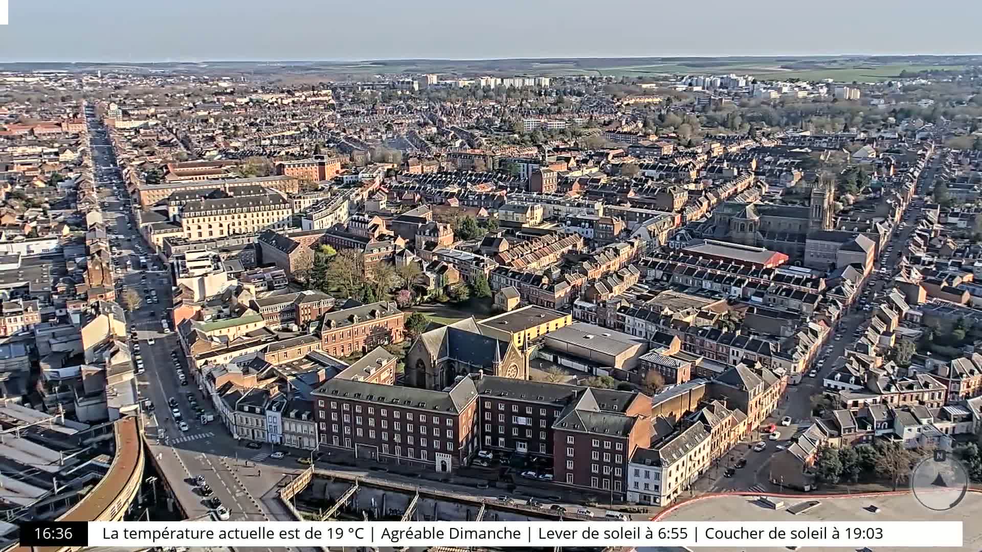 An aerial view shows a densely populated city on a sunny day, with a long, straight road cutting through the center.