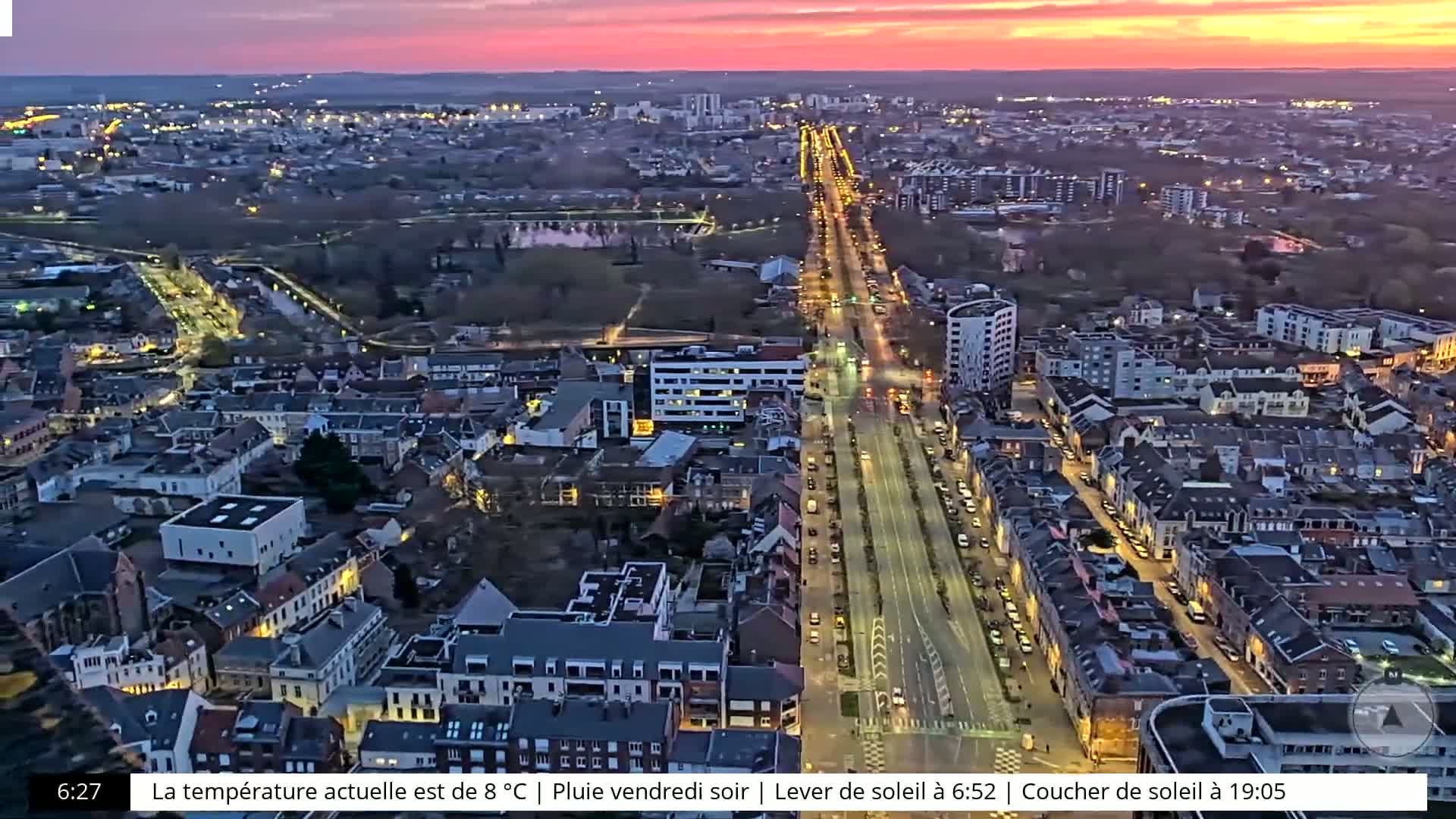 A nighttime aerial view of a city with a long, brightly lit street running through it, under a pink and purple sunset sky.
