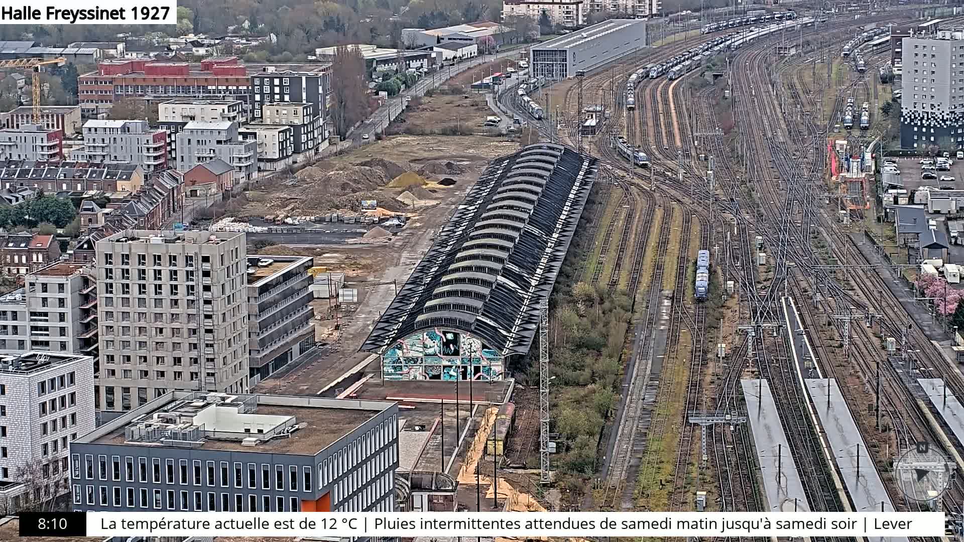 An aerial view shows a large railway yard with numerous tracks and trains, adjacent to a city with modern and older buildings under a cloudy sky.