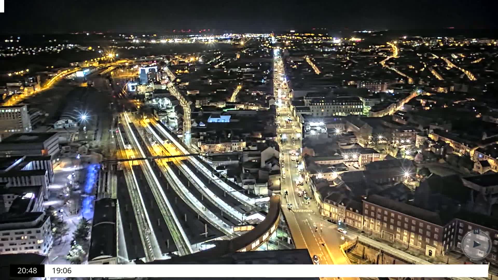 A nighttime aerial view of a city's illuminated streets, train station, and surrounding buildings.