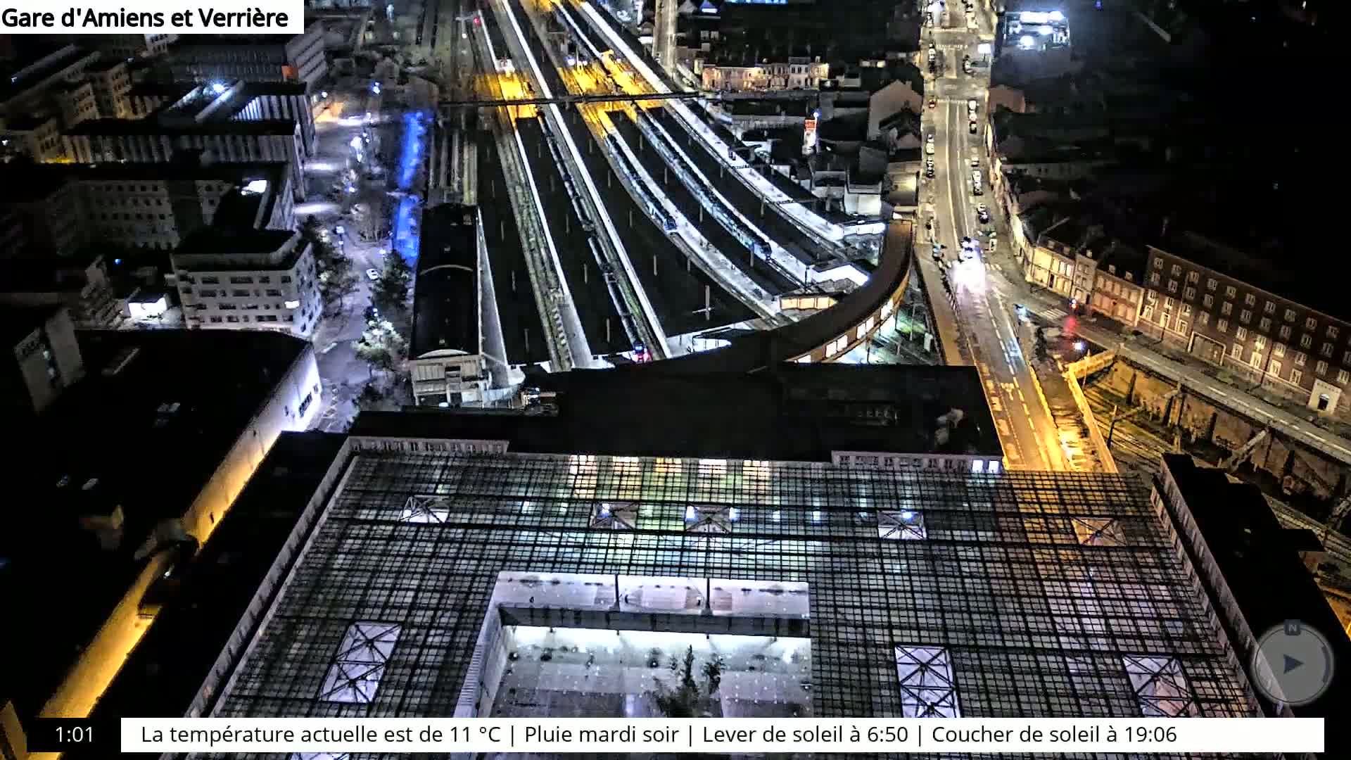 A nighttime aerial view of a train station and surrounding city streets shows several trains on illuminated tracks under a rainy sky.