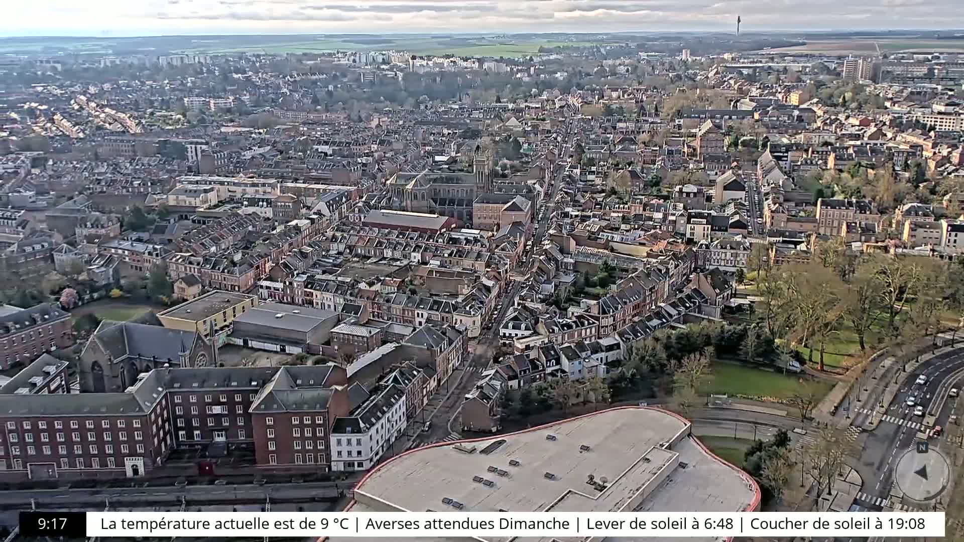 An aerial view of a densely populated city on a partly cloudy day, showing a mix of residential buildings and some green spaces.