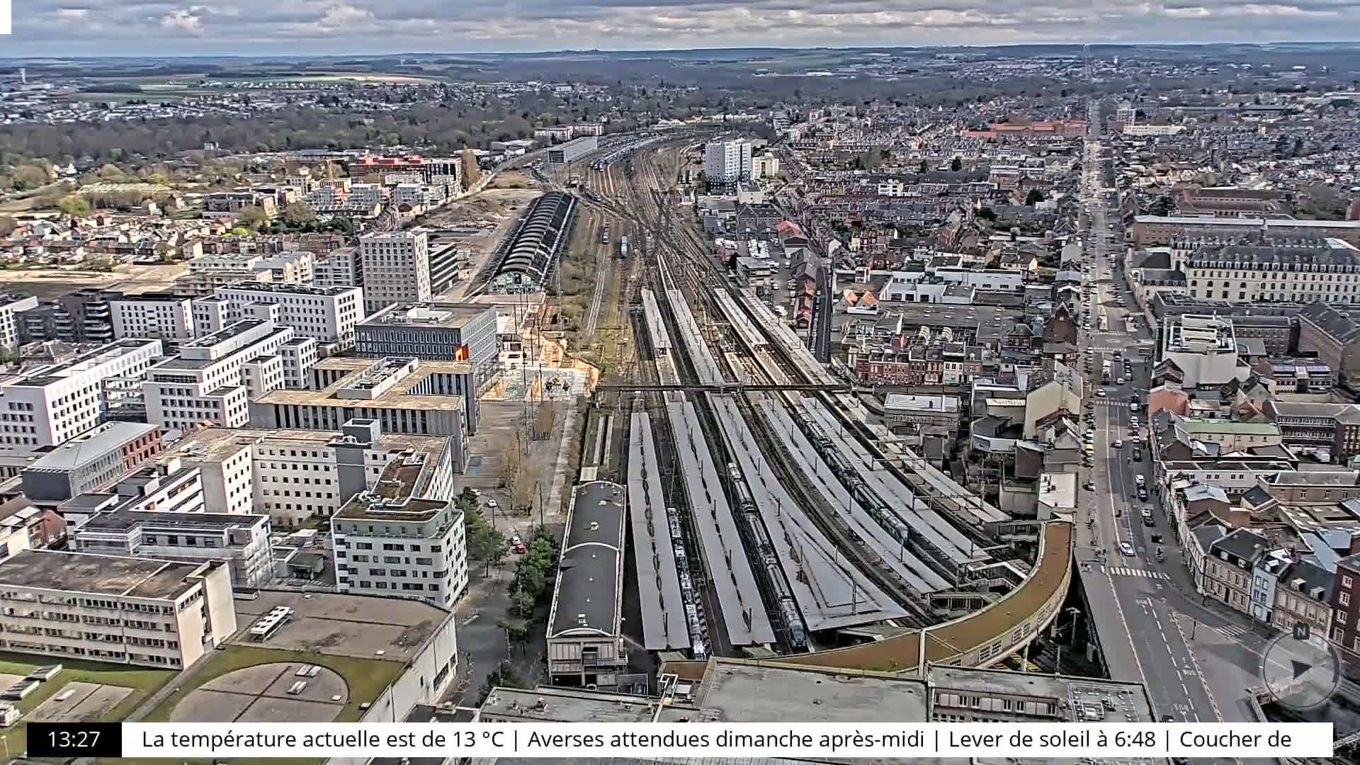 An aerial view shows a city with a large train station and many buildings under partly cloudy skies.