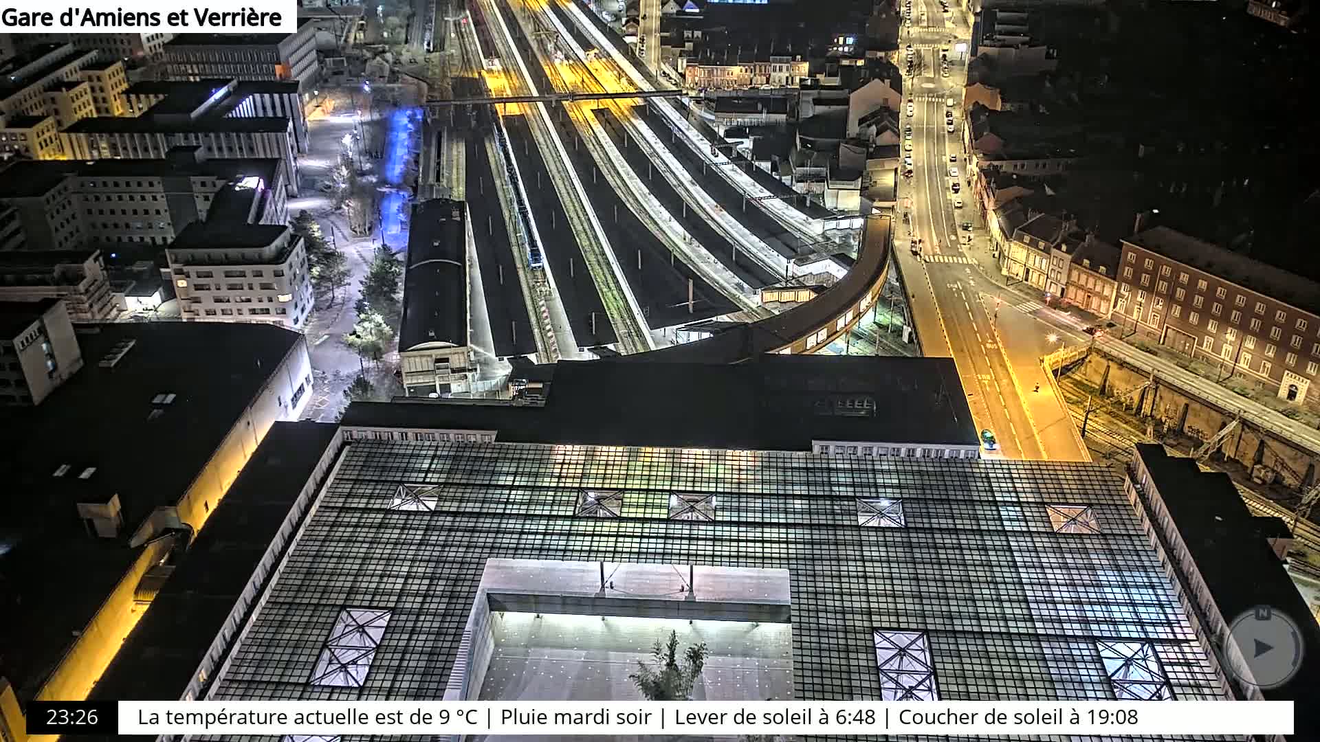 A nighttime aerial view of a train station and surrounding city streets under rainy conditions.