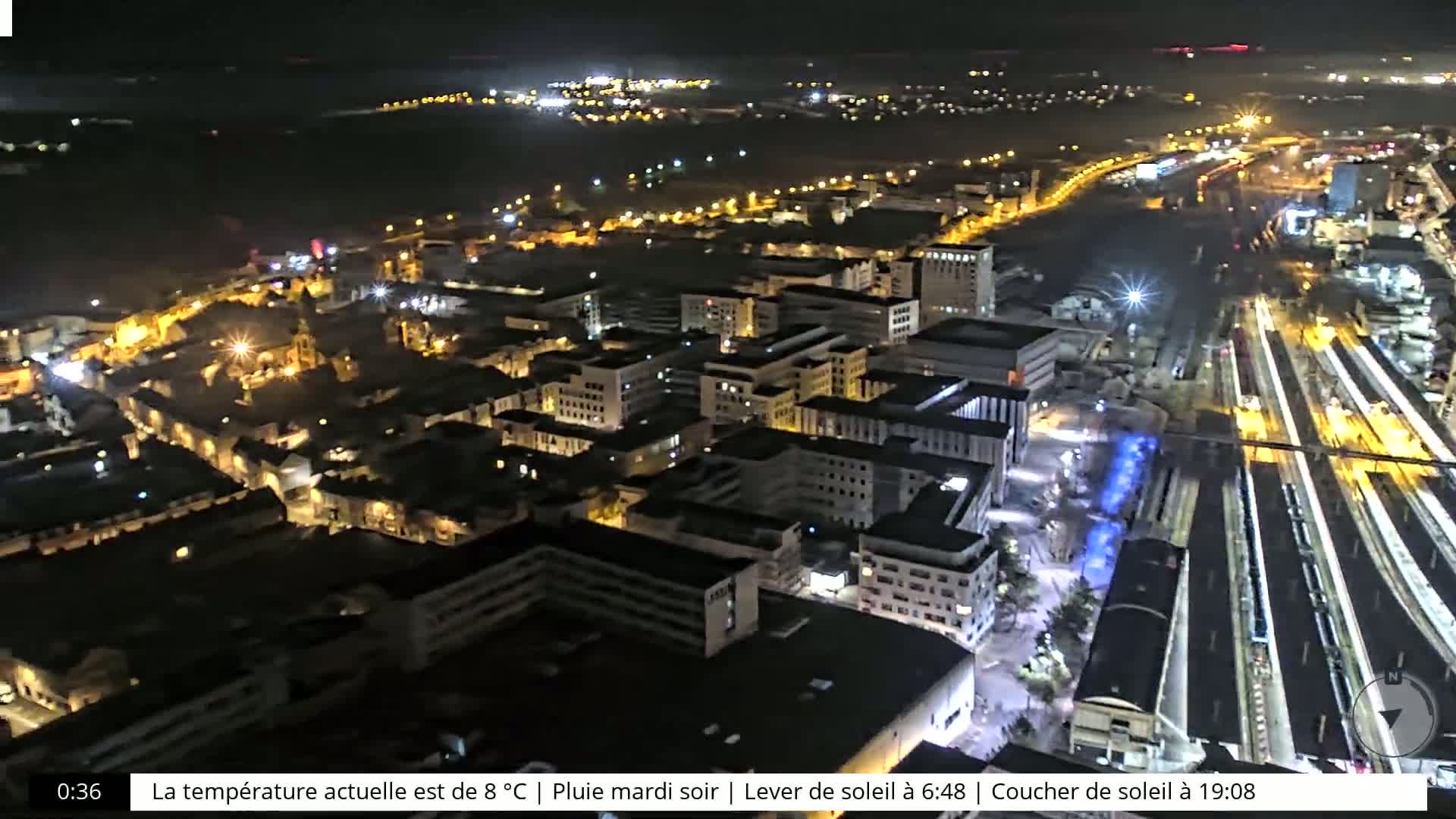 A nighttime aerial view of a city with rain, showing brightly lit buildings and a train station.