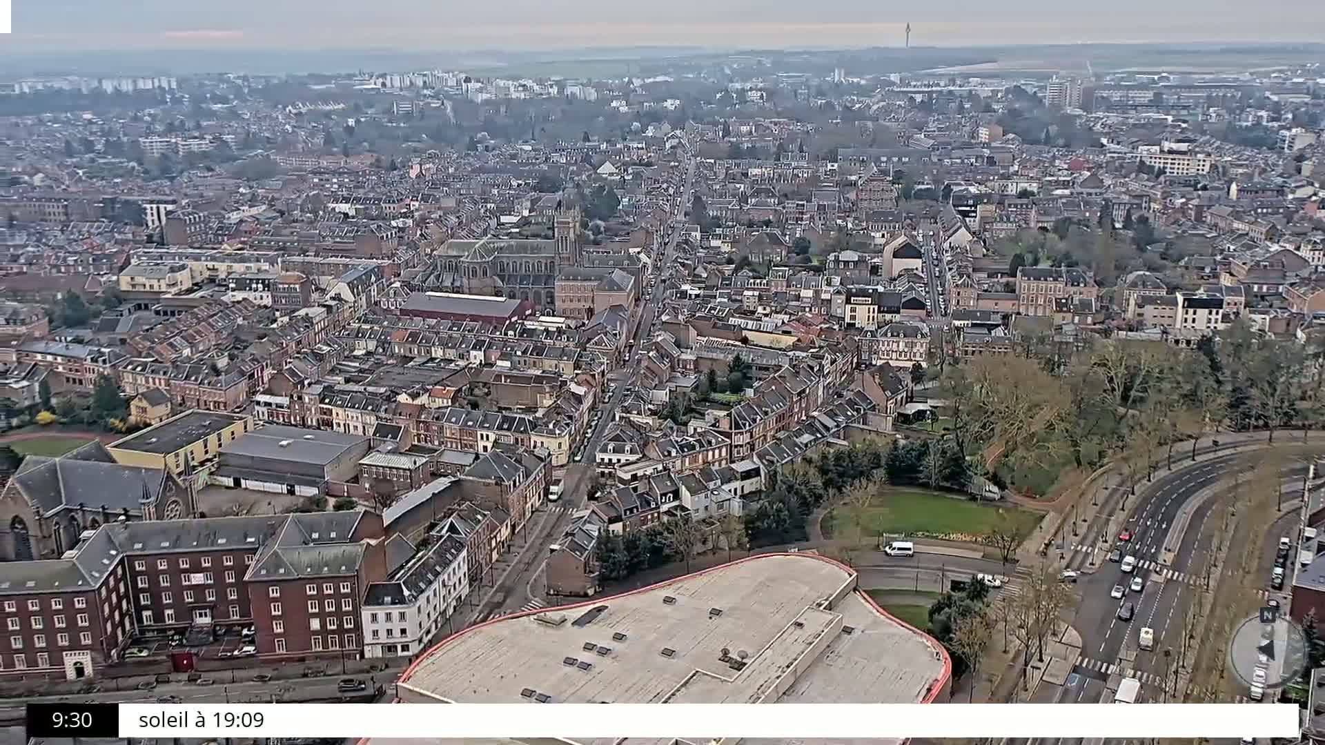 A high-angle, long shot of a city on an overcast day shows many closely packed buildings, roads, and a few trees.
