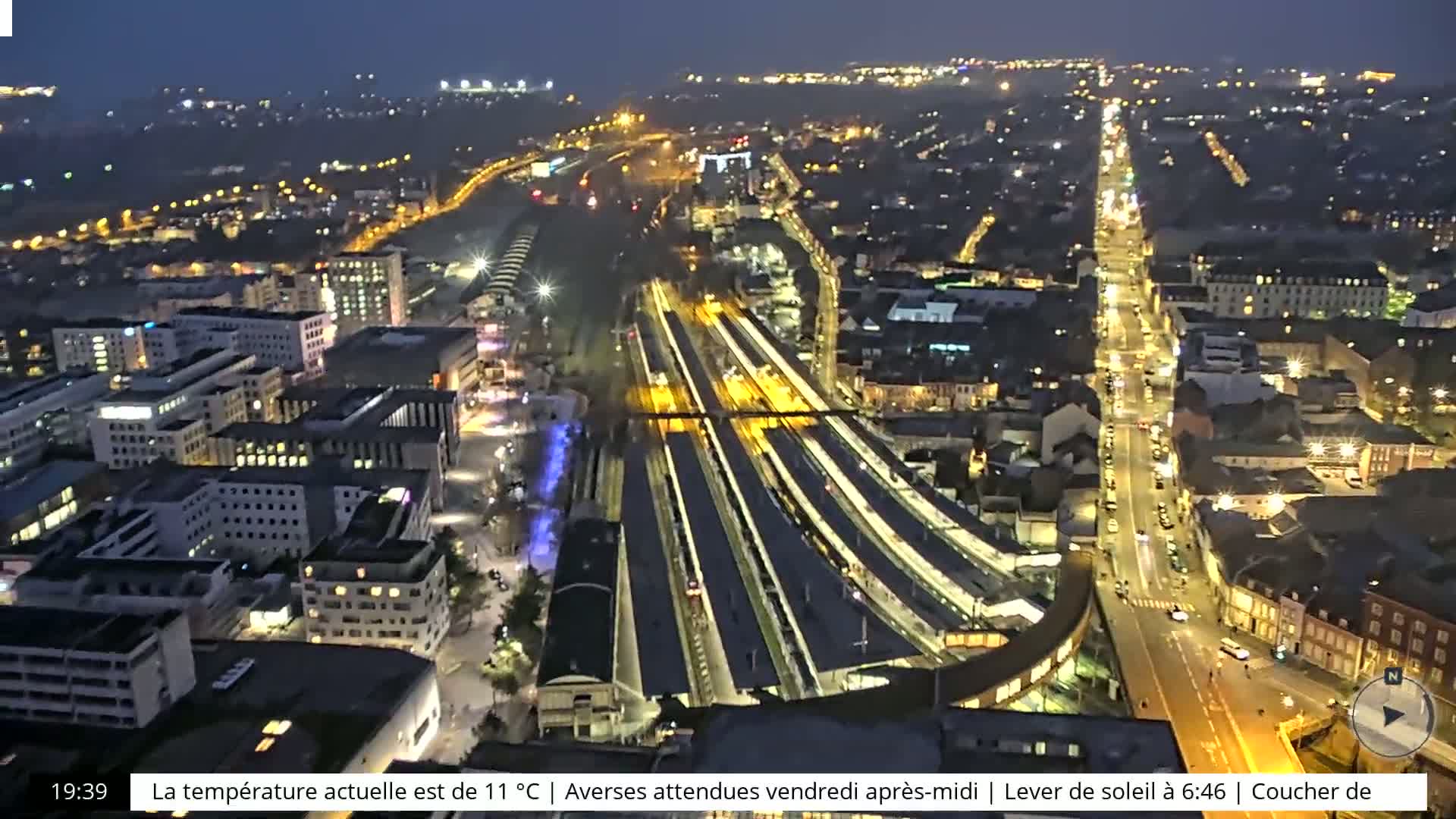 A nighttime aerial view of a city showing a train station and surrounding buildings illuminated with streetlights.