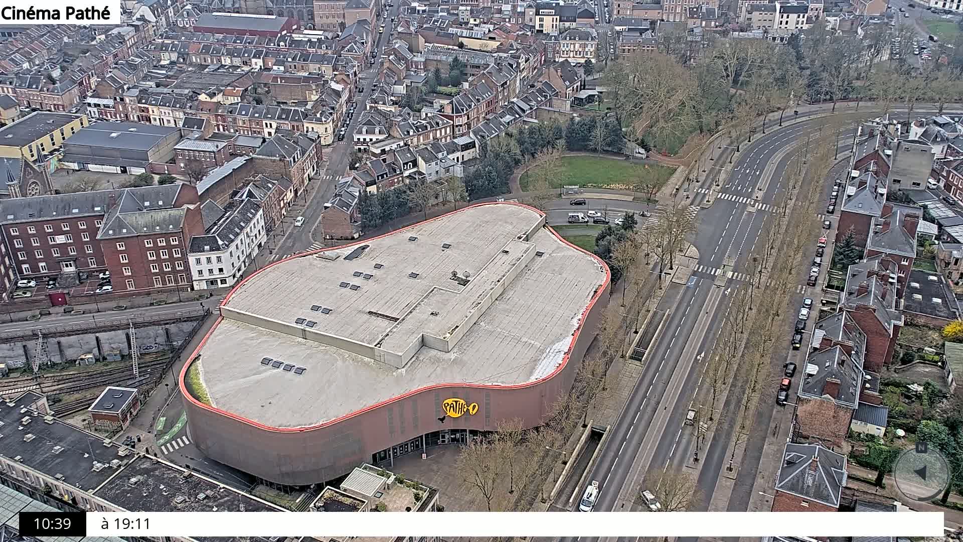 An aerial view shows a large, curved building next to a multi-lane road lined with trees and parked cars, all surrounded by a dense residential area on a mostly clear day.