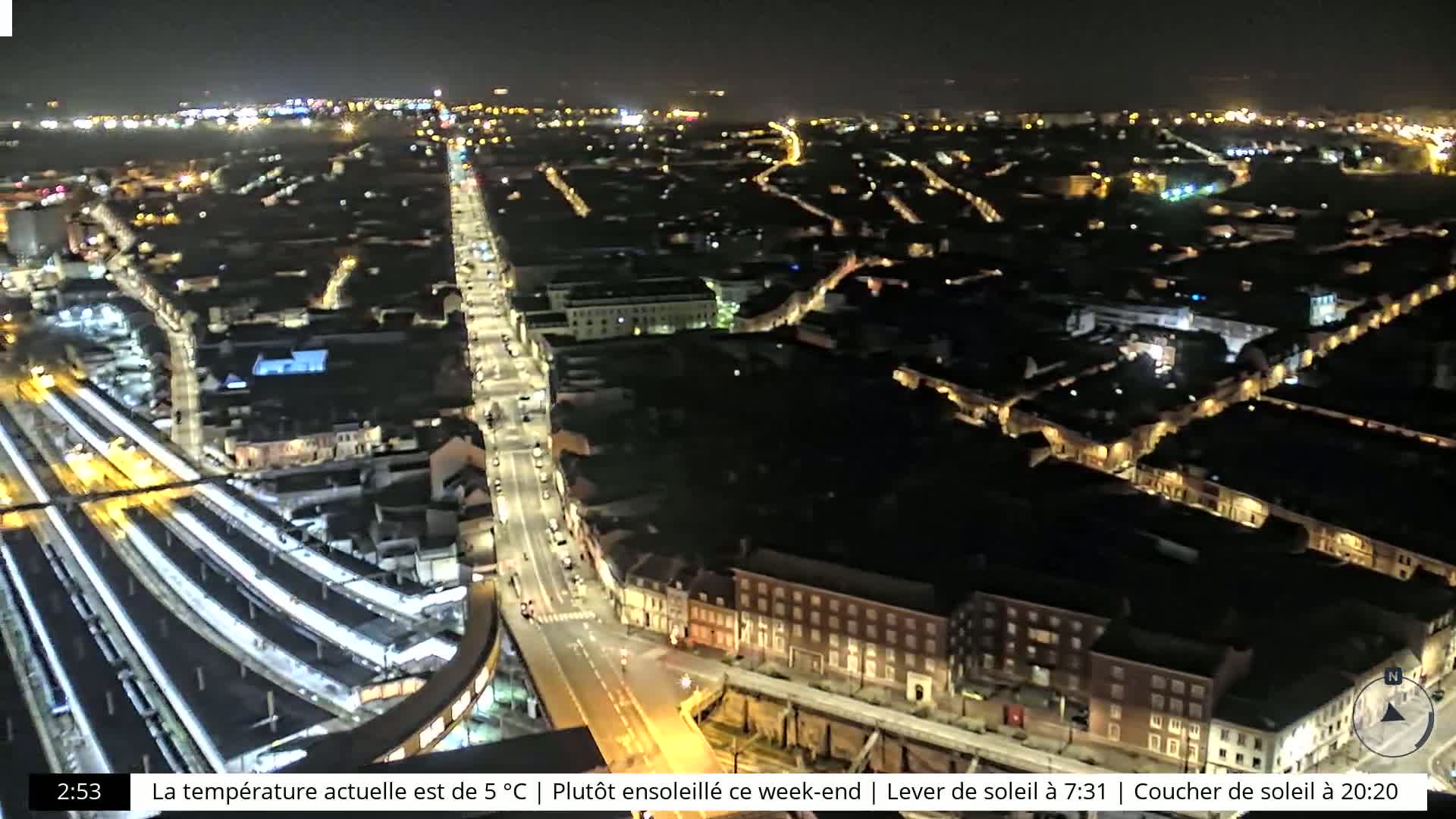 A nighttime aerial view of a city with illuminated streets and buildings, and a train station visible below.