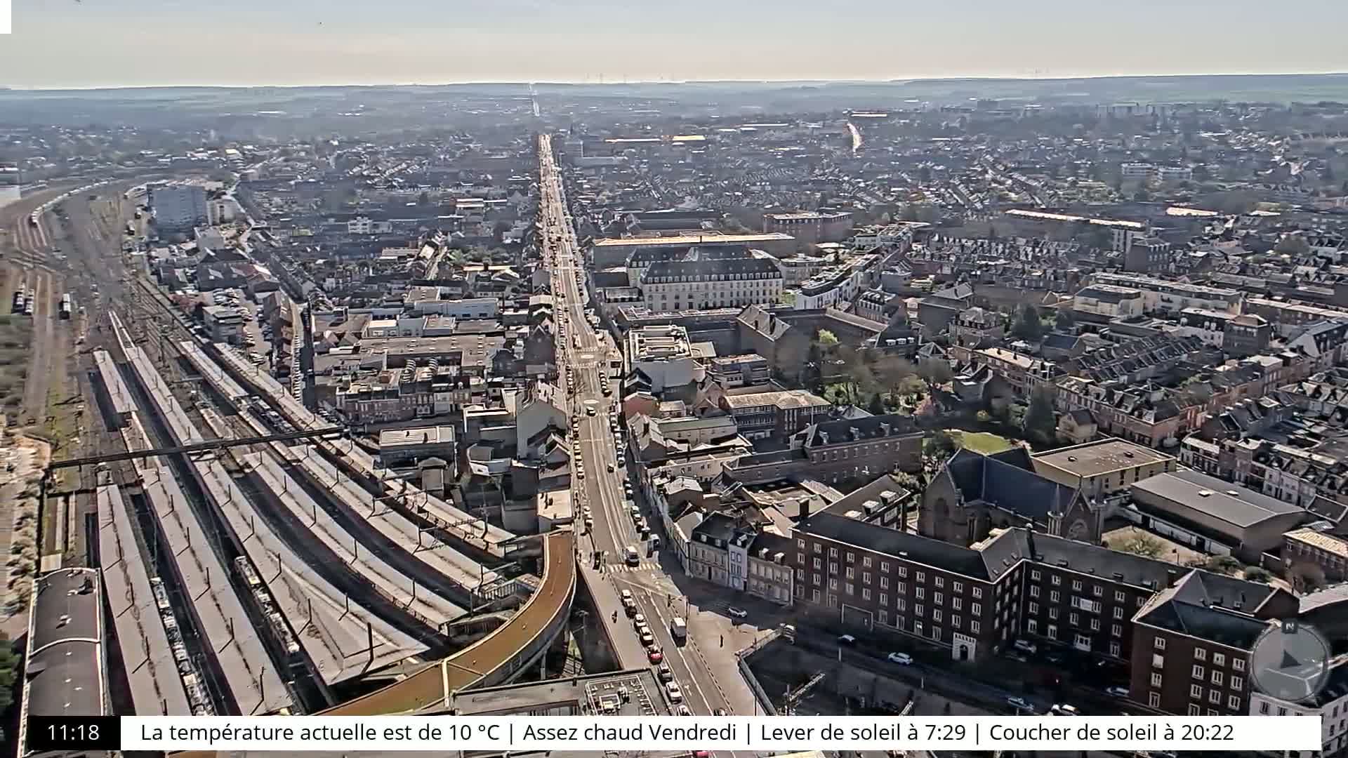 An aerial view of a city on a sunny day shows a long road with many parked cars, a train station with multiple tracks, and numerous buildings extending to the horizon.