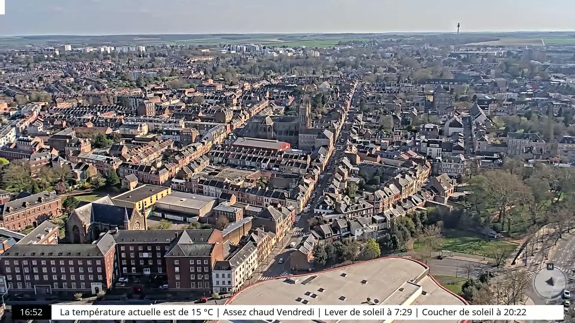 An aerial view of a city on a sunny day shows a dense collection of brick buildings surrounding a central thoroughfare, with a large church or cathedral visible in the middle.