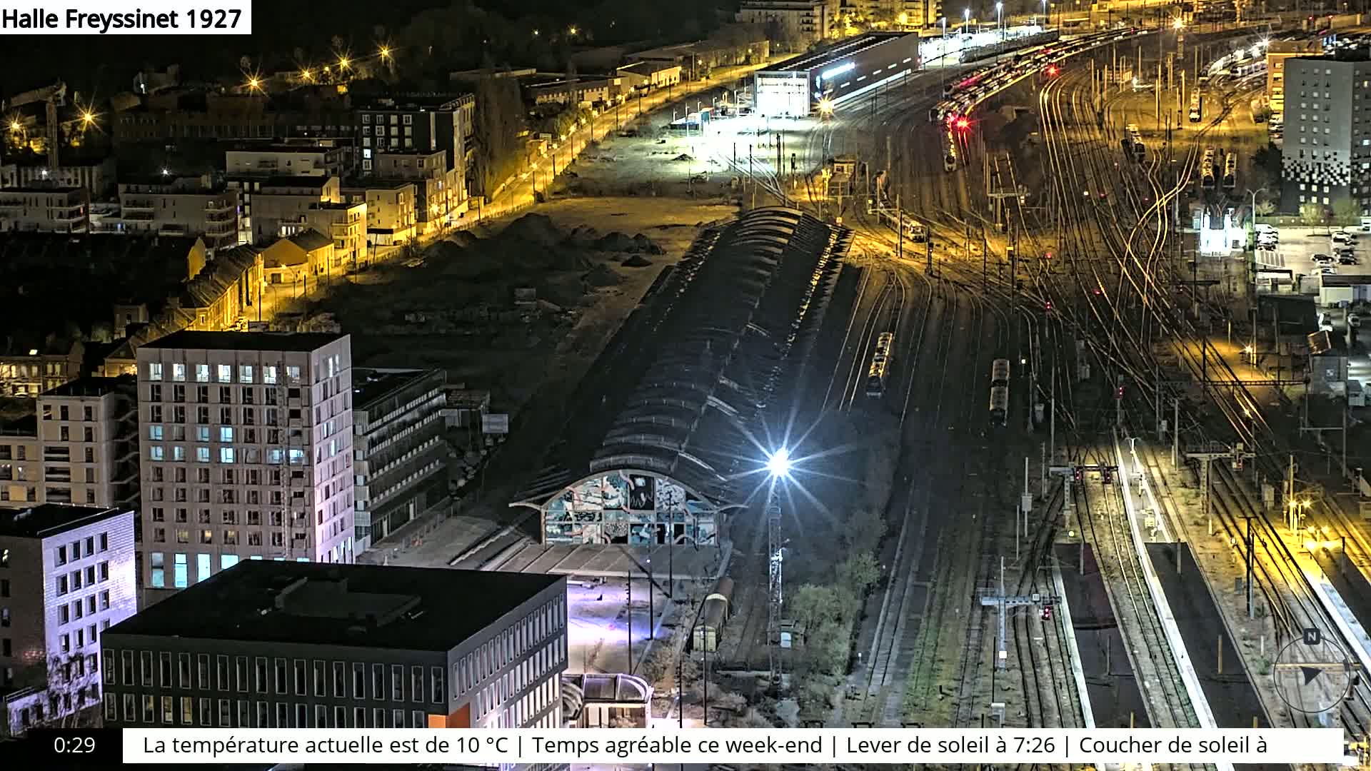 An aerial nighttime view of a train station and surrounding city buildings, illuminated by streetlights and building lights.