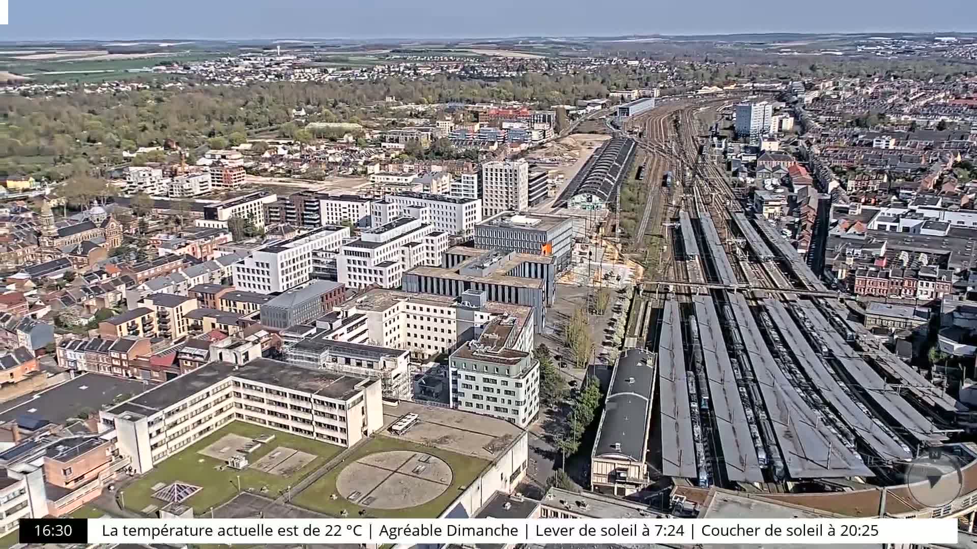 An aerial view of a city on a sunny day shows a large train station with many trains, surrounded by buildings of varying heights and styles, some under construction.