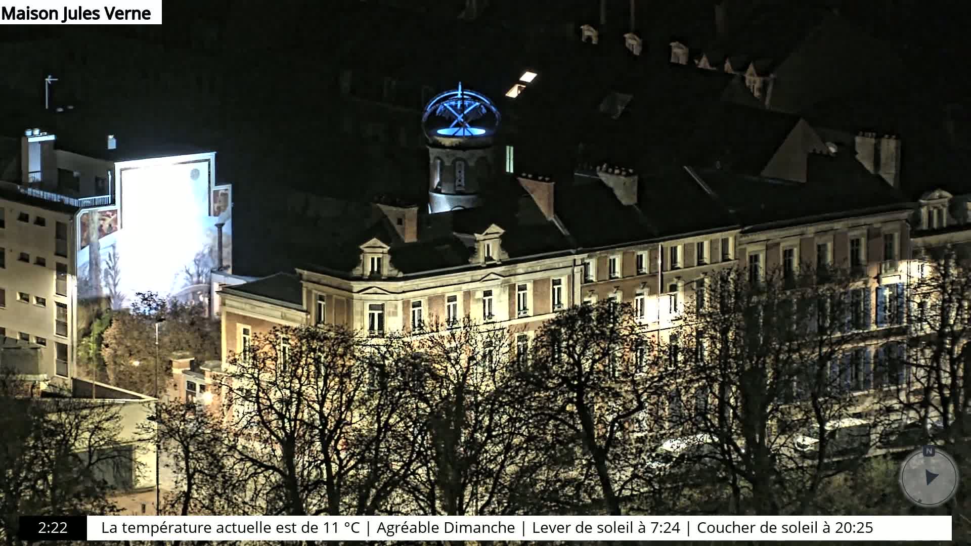 A nighttime view of buildings with a blue-lit dome, seen from above, surrounded by bare trees under a clear, dark sky.