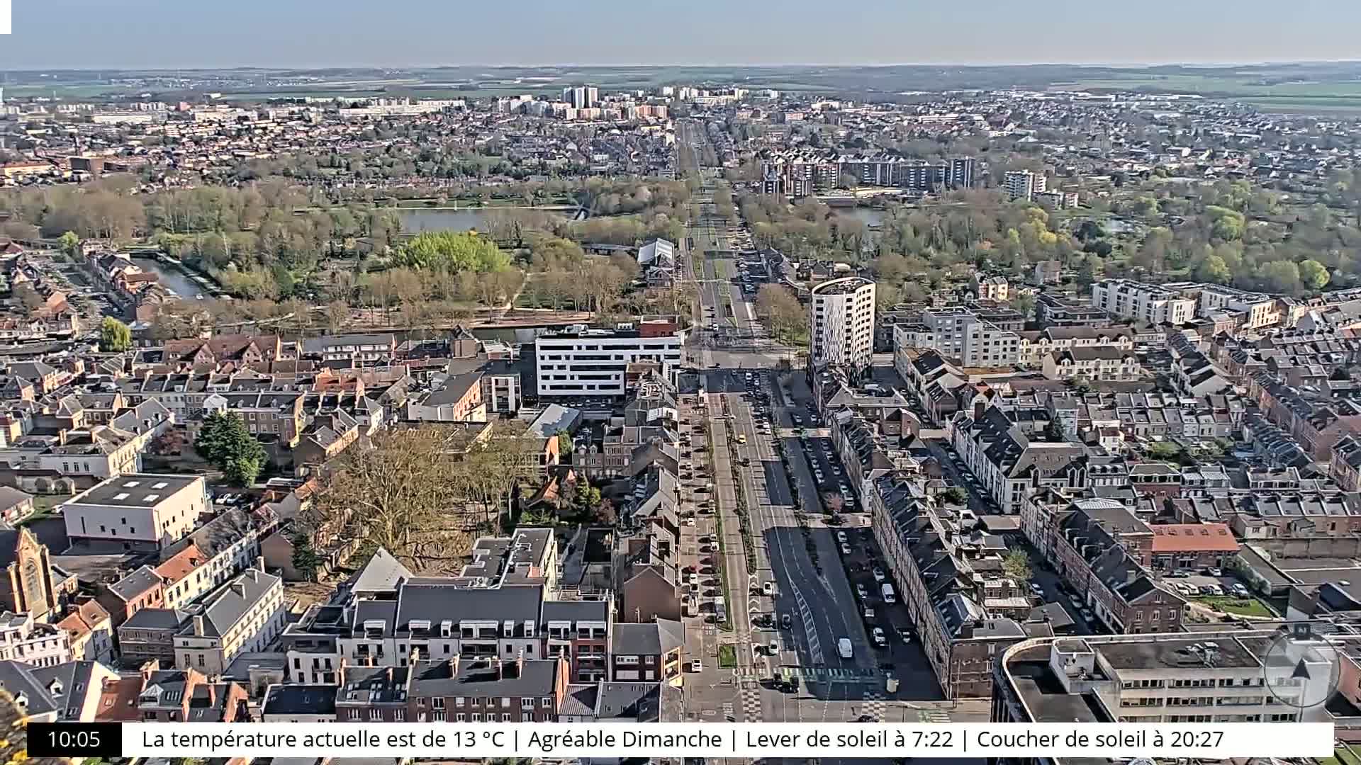 A sunny aerial view of a town shows a long road lined with buildings and trees, with a body of water and surrounding greenery visible in the distance.