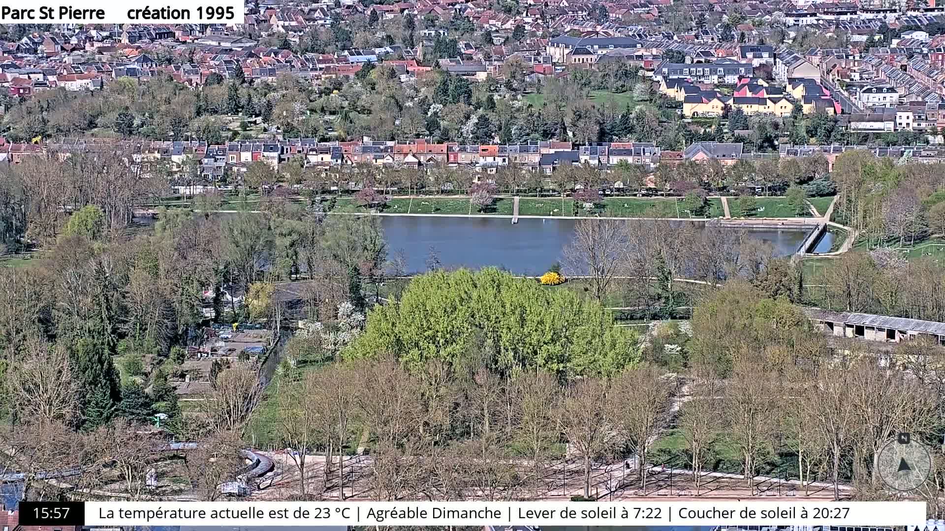 An aerial view shows a park with a pond, surrounded by trees and residential buildings under a sunny sky.