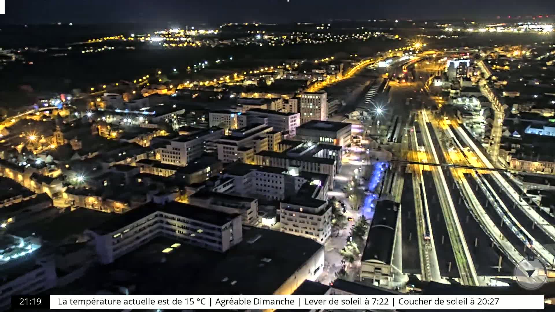 A nighttime aerial view of a city with brightly lit buildings and train tracks, under a clear sky.