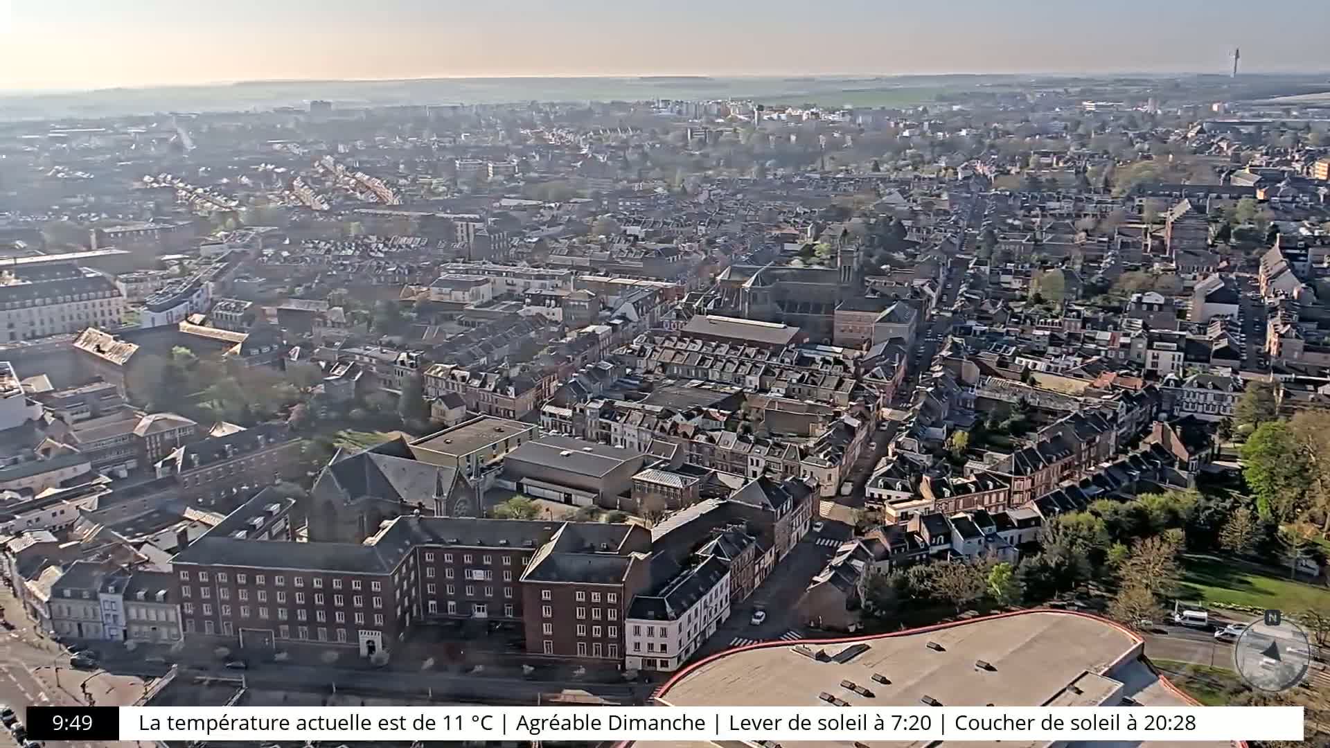 A high-angle, long shot of a city on a sunny day shows many closely packed buildings and streets.