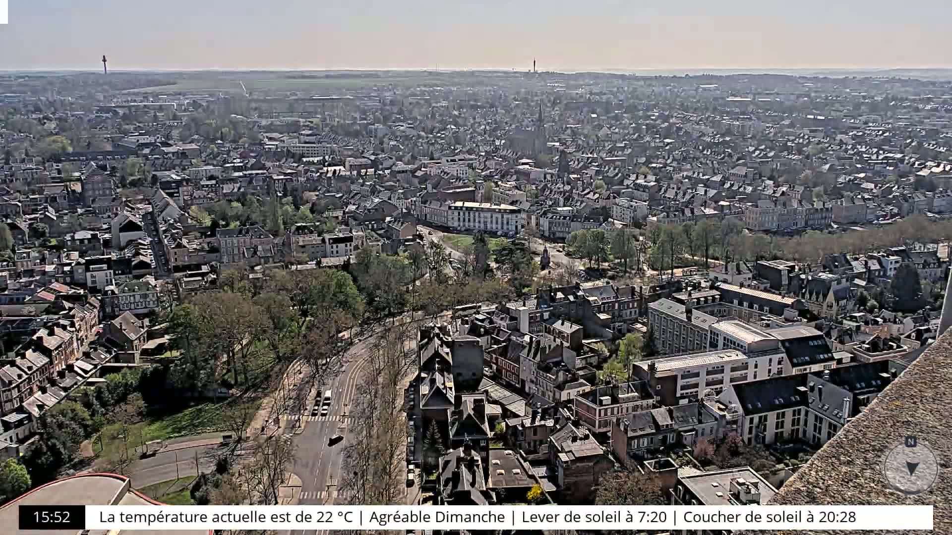 A high-angle, long shot of a city on a sunny day shows many closely-packed buildings and tree-lined streets.