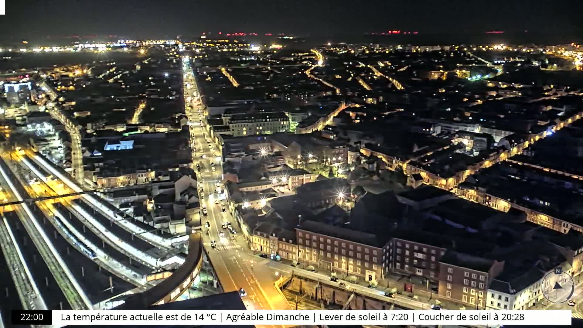 A nighttime aerial view of a city with brightly lit streets and train tracks, under a clear sky.