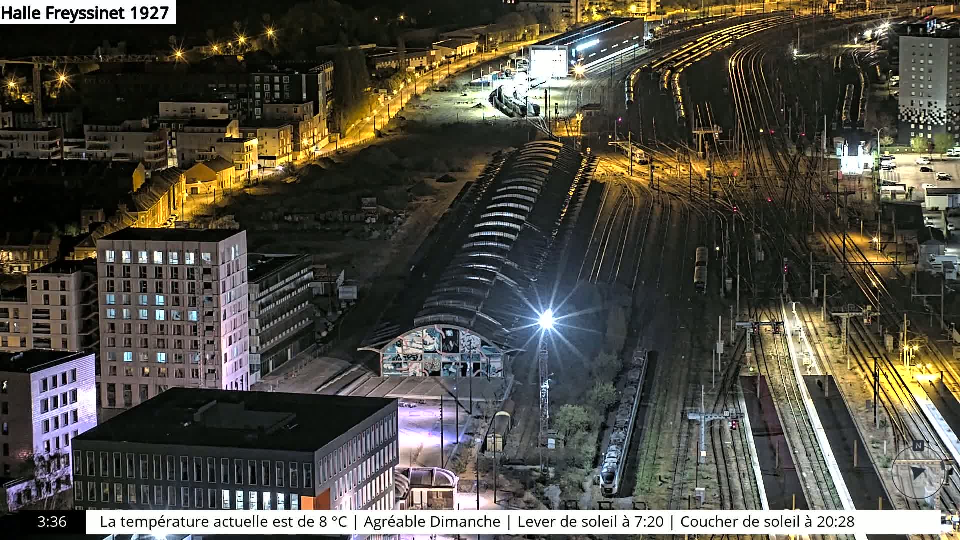 A nighttime aerial view of a train station and surrounding city, showing a network of train tracks, buildings with illuminated windows, and a large arched structure.