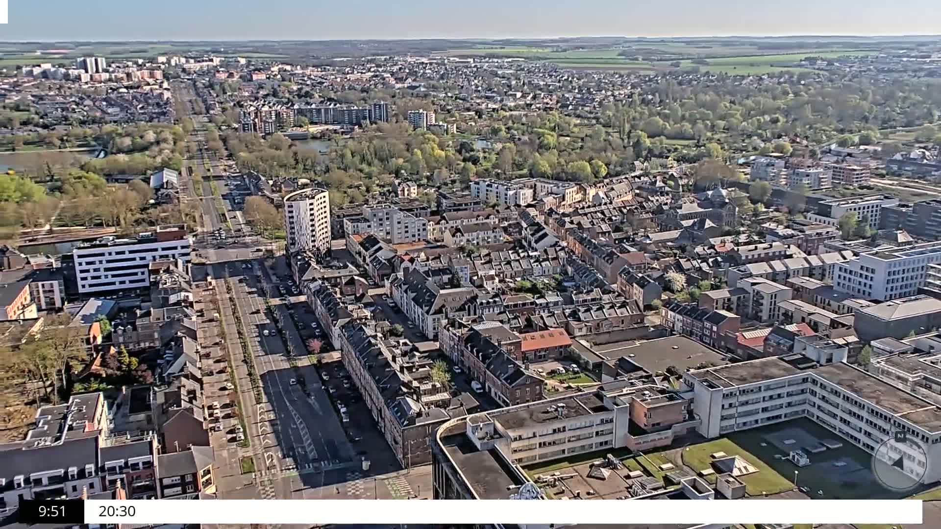 An aerial view shows a densely populated residential area on a sunny day, with a long road running through the center and green spaces interspersed among the buildings.