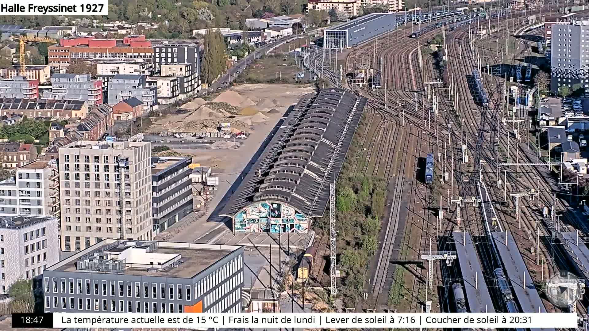 An aerial view shows a large train yard with many tracks and trains, adjacent to a city with buildings under construction and a graffiti-covered structure.