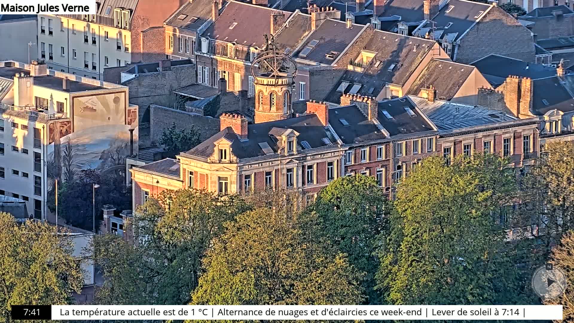 An aerial view shows a city block of buildings with red-brick facades, lush green trees, and a building with a unique spherical structure on its roof, all bathed in the golden light of sunrise.