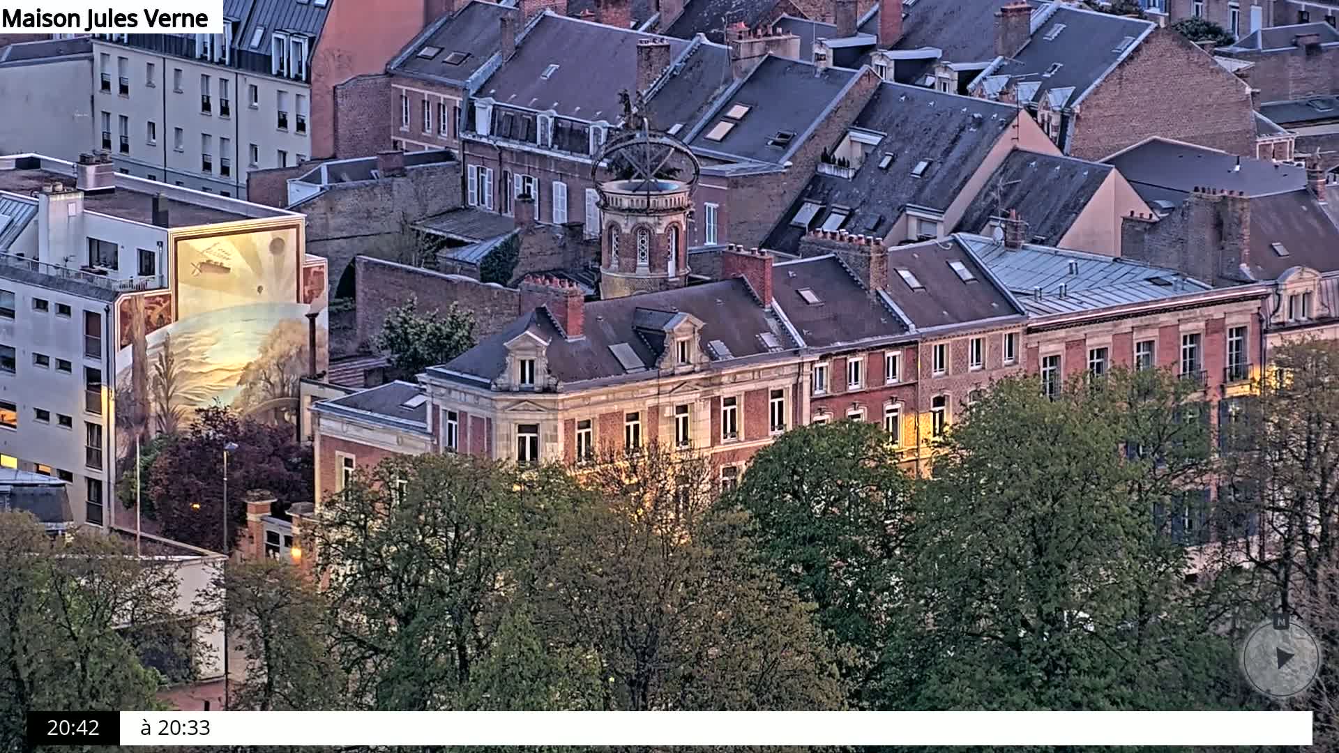 An aerial view at dusk shows a large building with a rooftop structure, surrounded by other buildings and green trees.