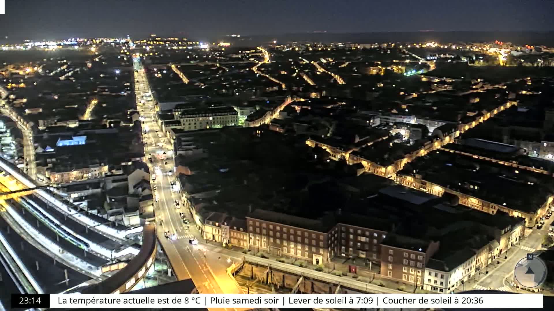 A nighttime aerial view of a city with illuminated streets and buildings, under rainy conditions.