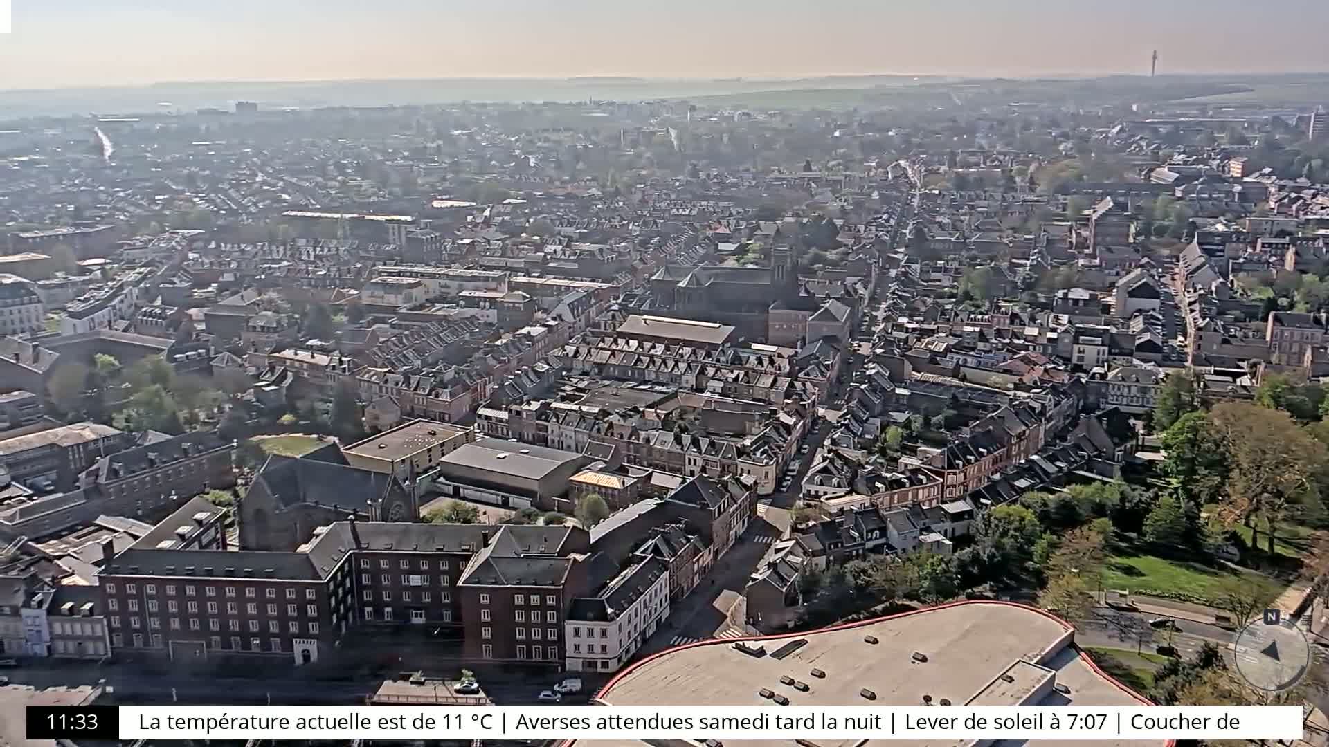 A high-angle, long shot on a sunny day shows a sprawling city with many buildings and streets, featuring a mix of architectural styles and green spaces.