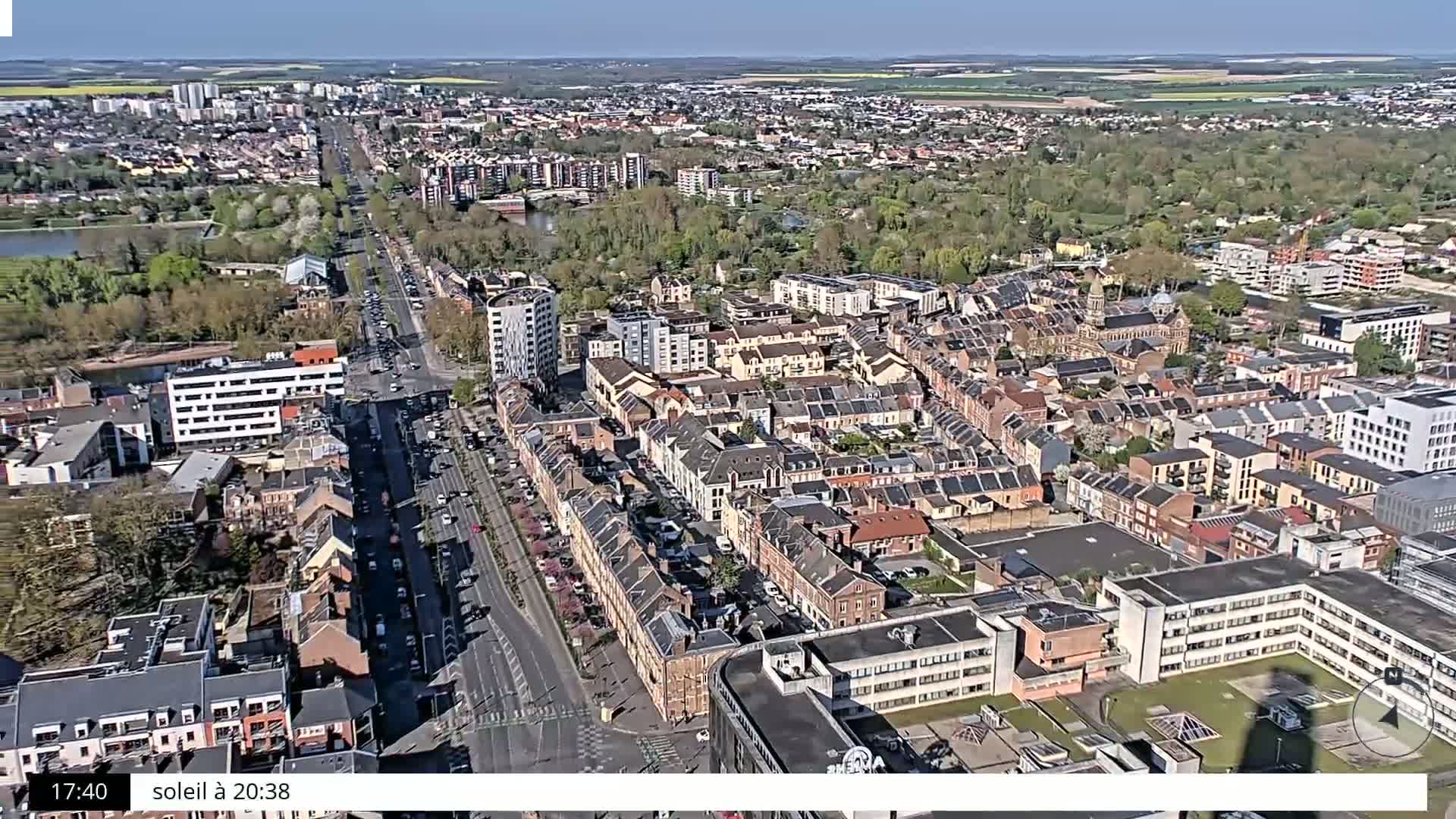 An aerial view of a city on a sunny day shows a long road with cars driving through a dense residential area with many buildings.